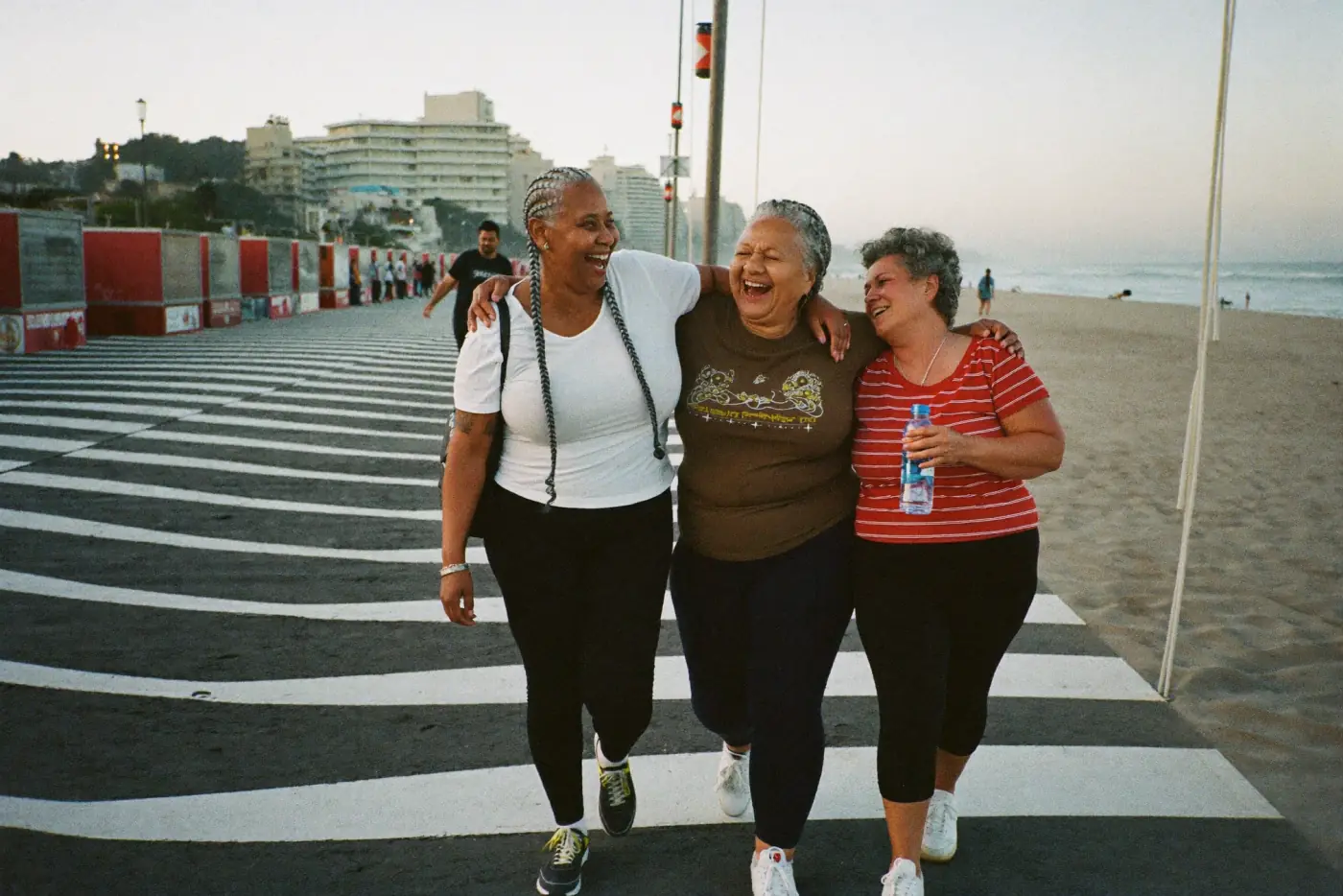 Três amigas brasileiras com mais de 50 anos caminhando abraçadas e rindo no calçadão da praia
