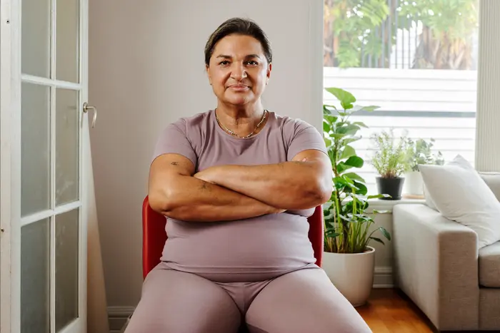 Mulher de uns 55 anos sentada numa cadeira na sala, de braços cruzados, pronta para o exercício de sentar e levantar