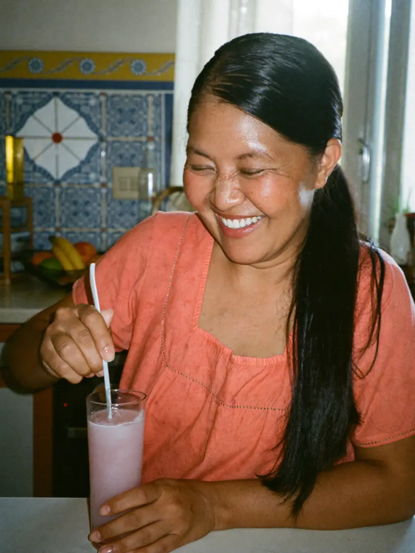 Mulher de cerca de 45 anos sorrindo enquanto mexe uma bebida rosa num copo, na cozinha